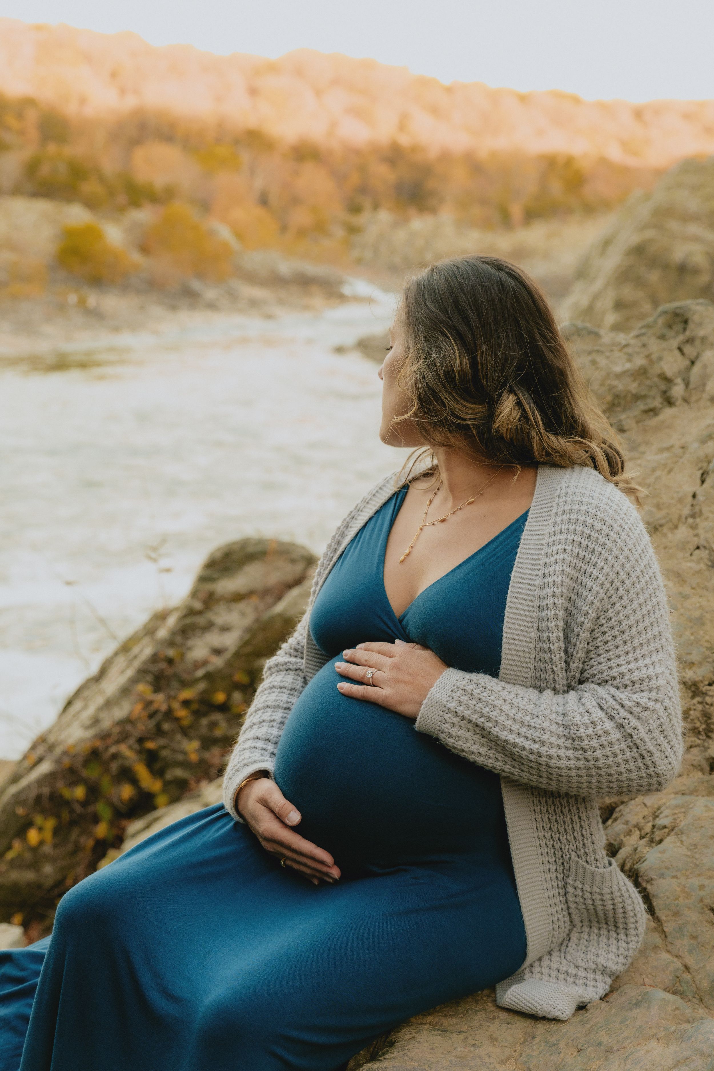 Expectant mother in a blue dress and cardigan sits peacefully on coastal rocks.