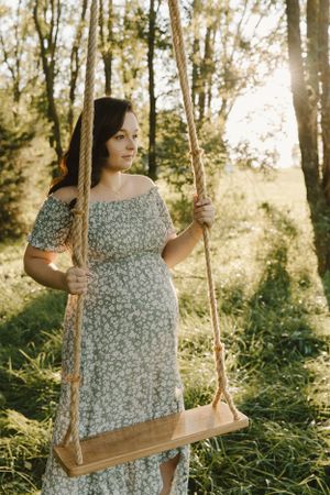 Rustic maternity portrait on a wooden swing in a sunlit forest clearing.