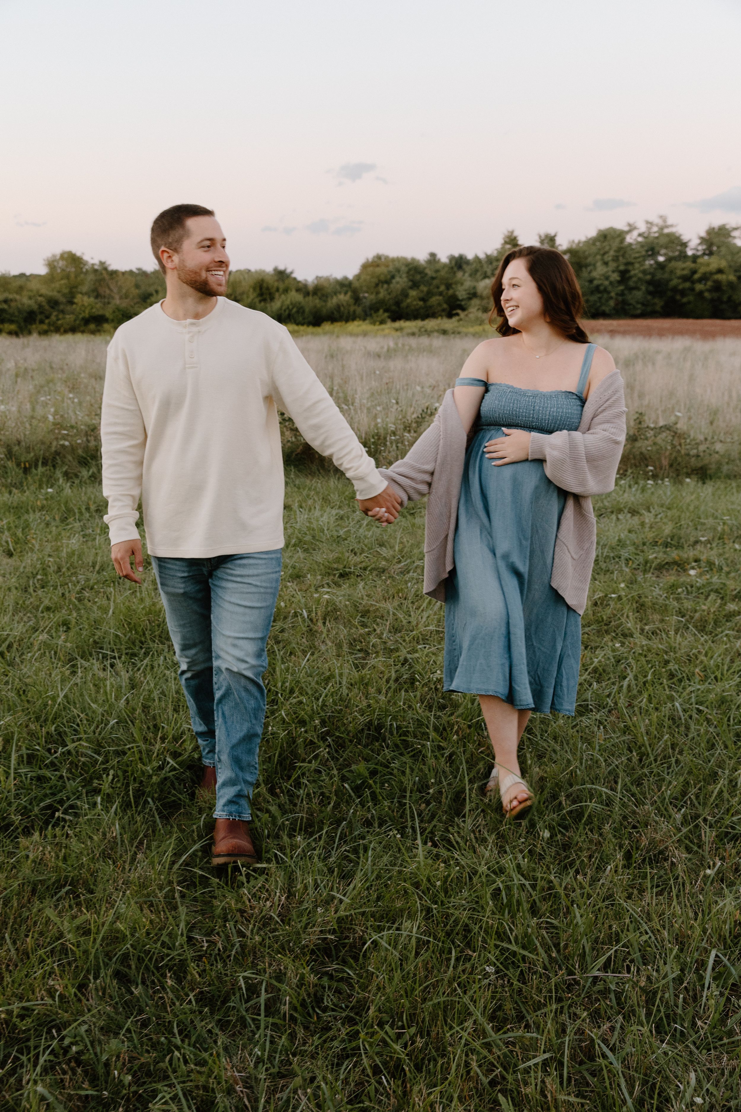 A couple walking hand in hand through a grassy field at sunset.
