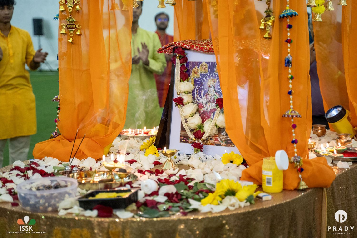 A decorated Hindu shrine with orange drapes, flower petals, and religious offerings during a festive celebration.