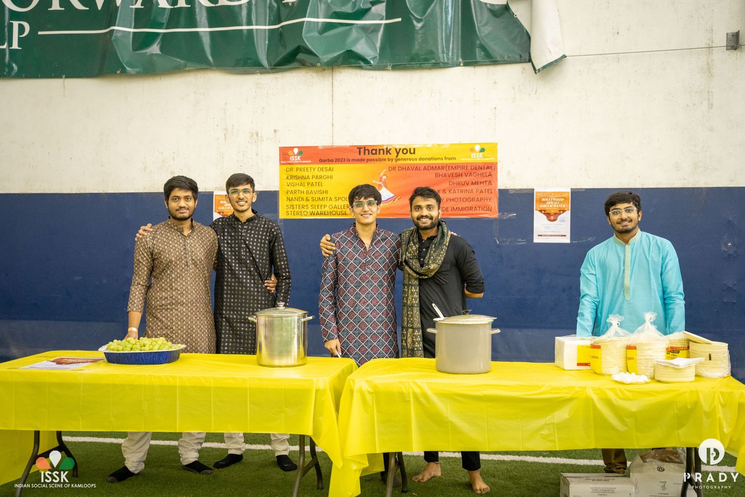 People in traditional Indian attire standing behind a table with yellow tablecloth at an indoor event.