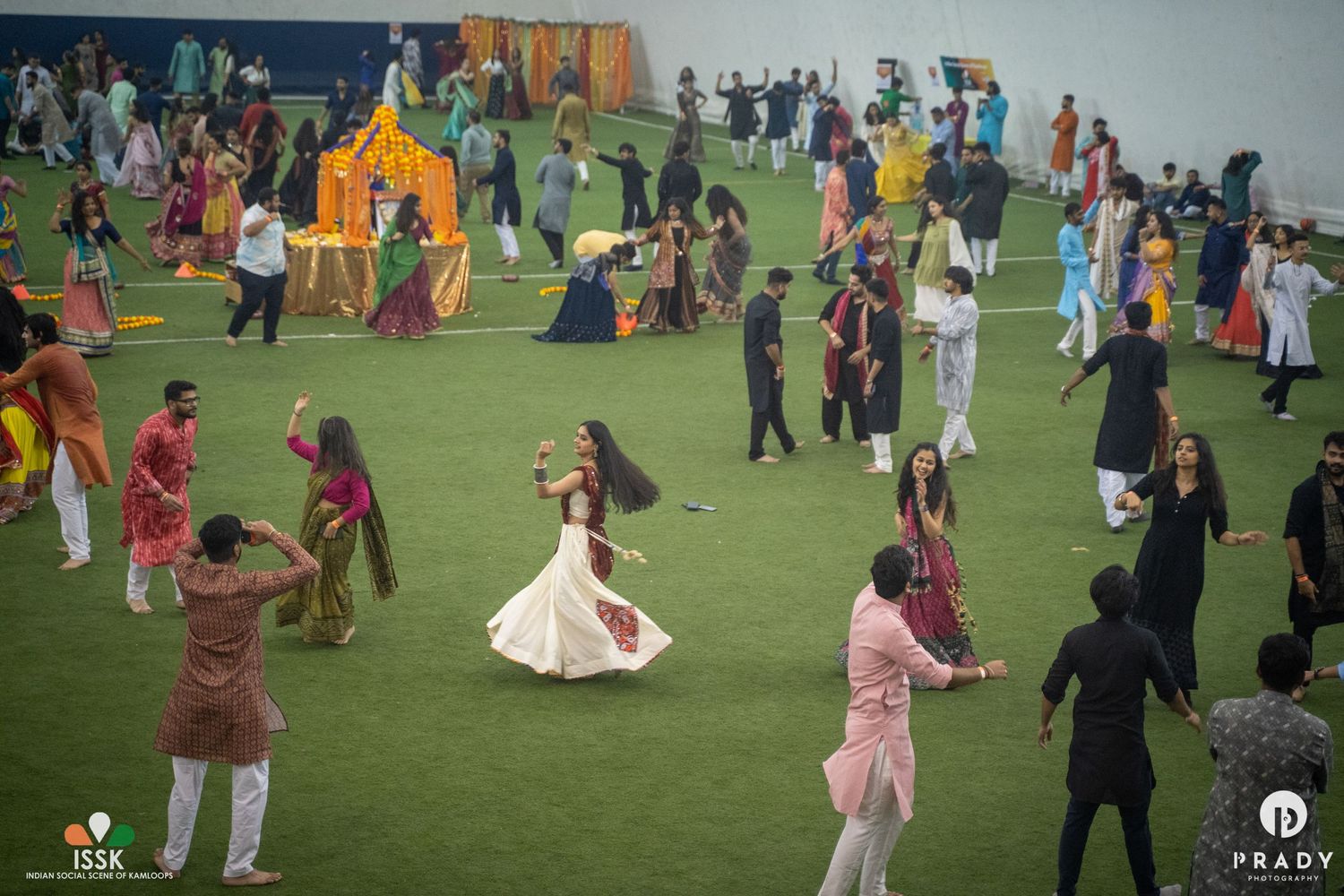 Large group of people dancing and celebrating on a green indoor field with colorful decorations.