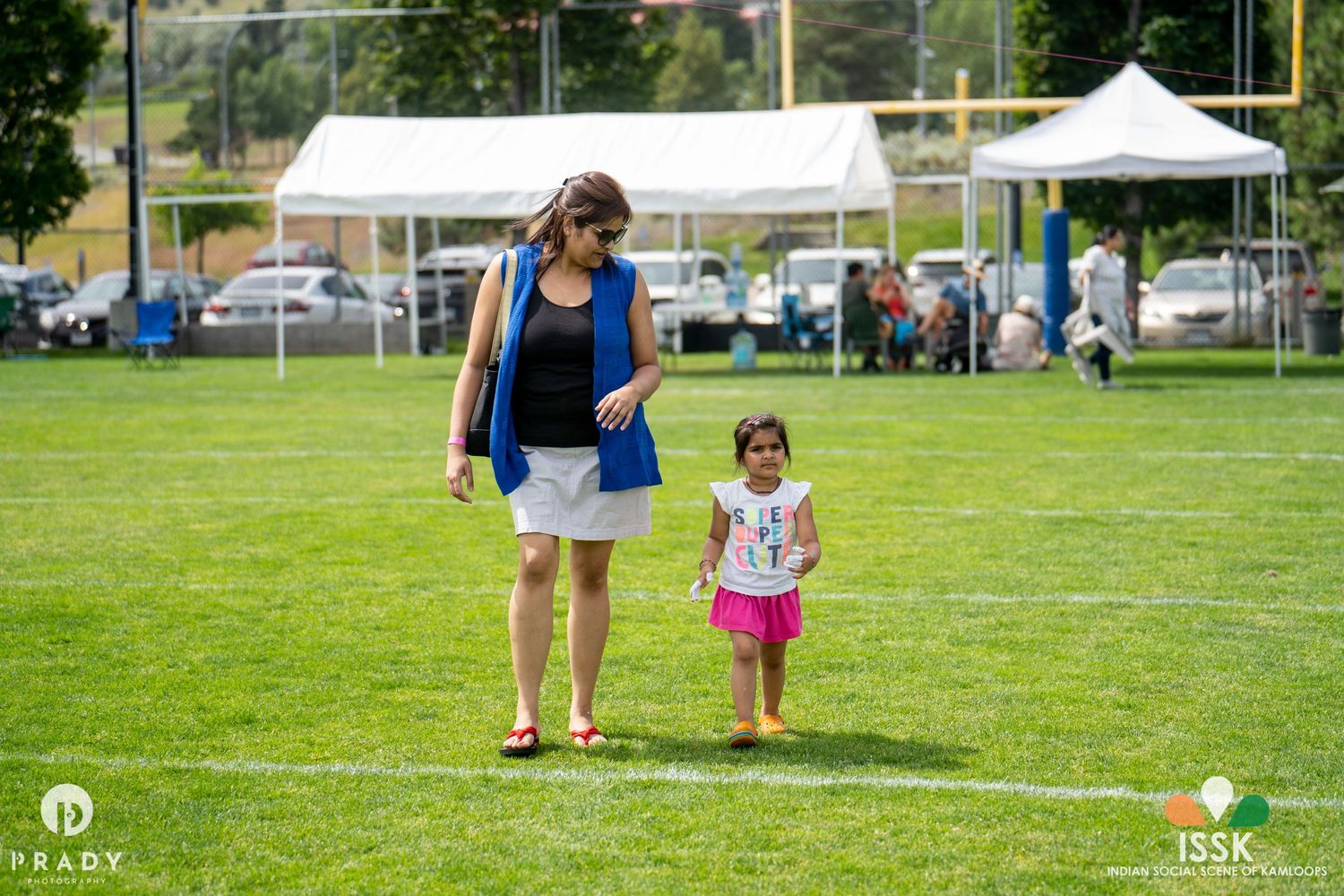 Two people walking together on a green lawn with white tents in the background.
