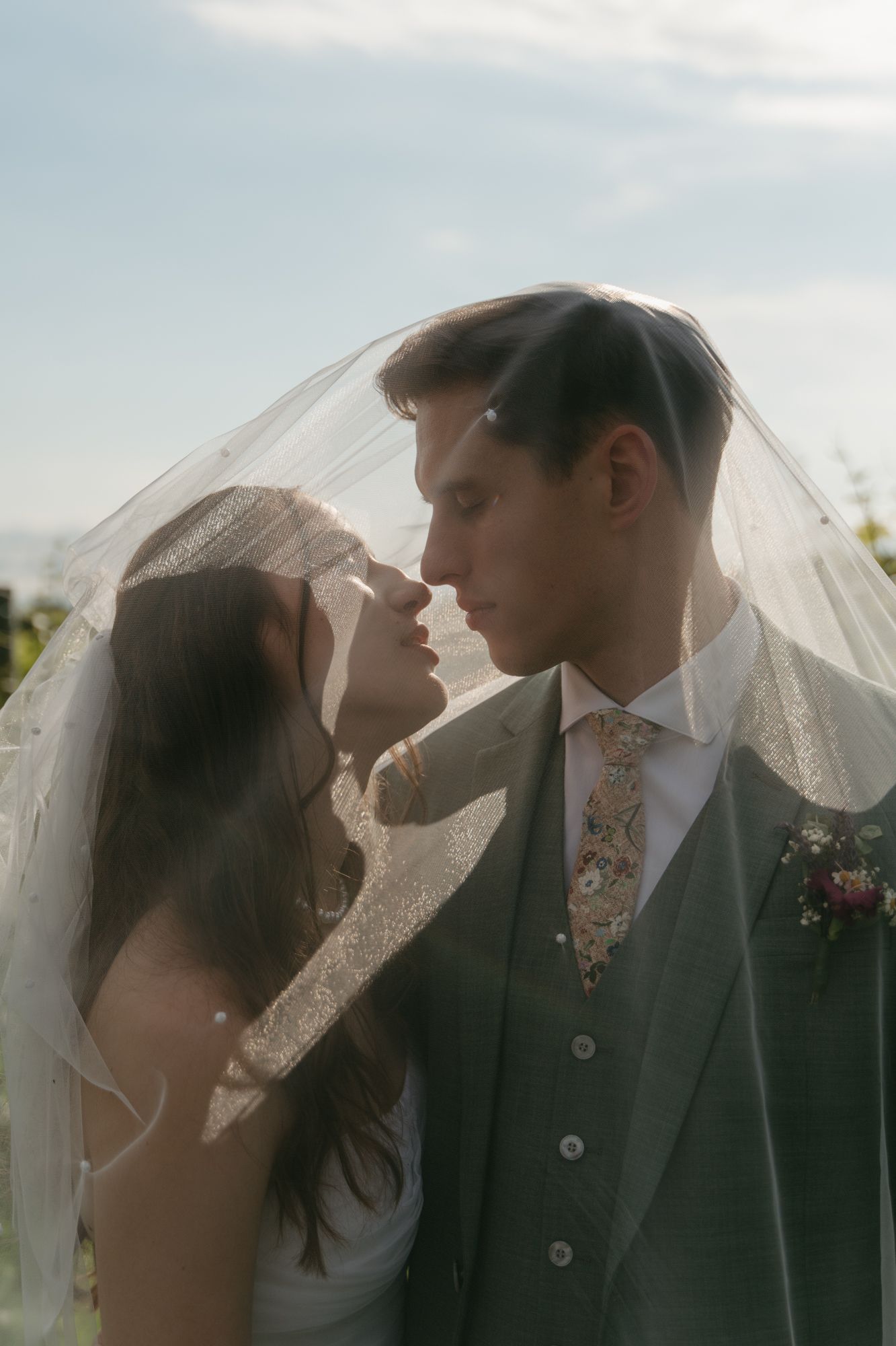 Romantic wedding portrait with couple under flowing veil against soft sky.