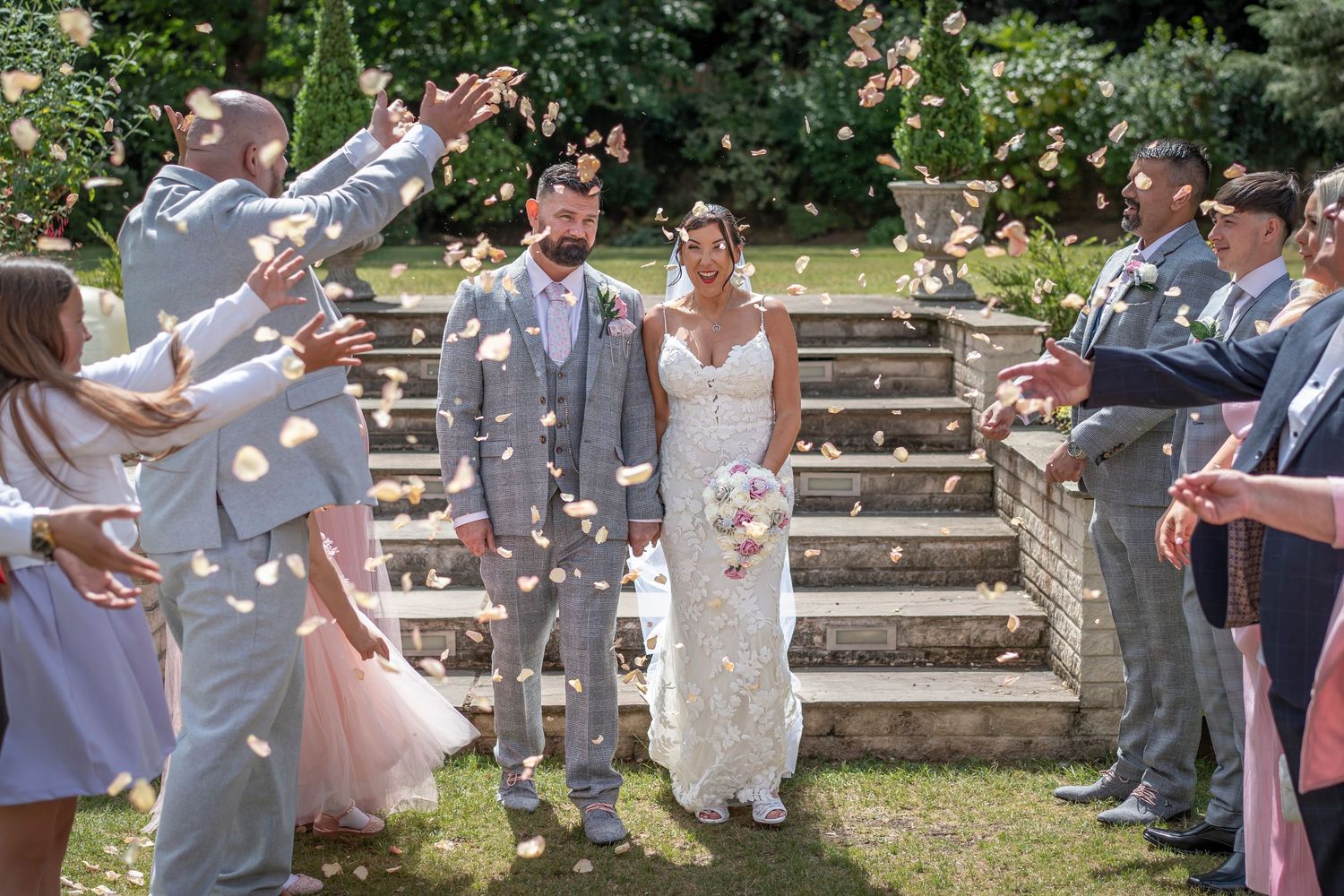 Wedding party throws flower petals on outdoor stone steps during summer celebration.