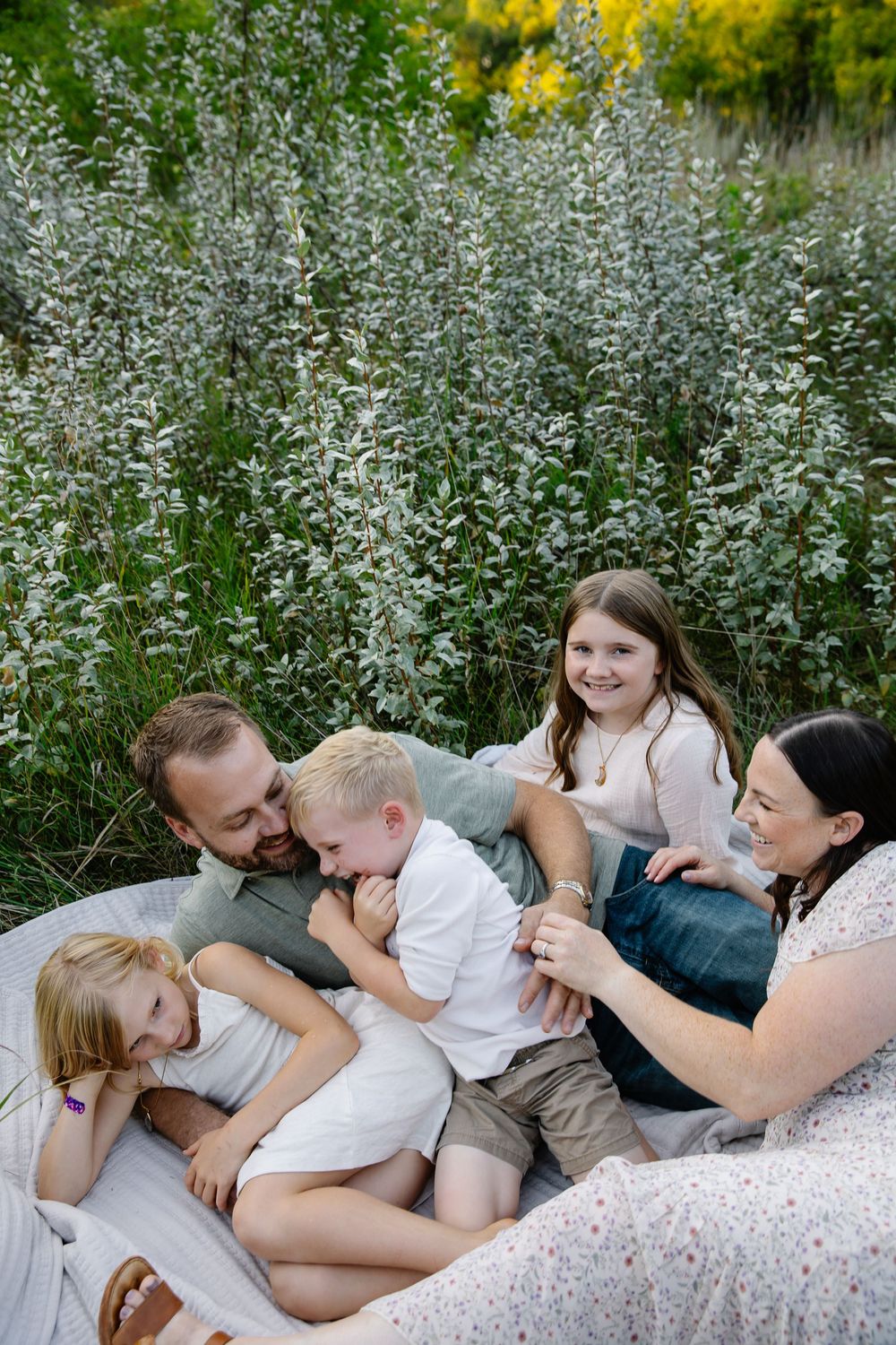 Family lying together on blanket outdoors surrounded by tall flowering plants.