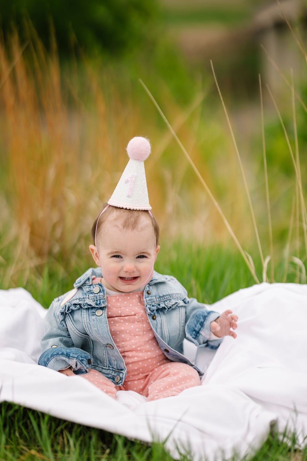 Baby portrait session outdoors with infant wearing denim jacket and party hat on white blanket.