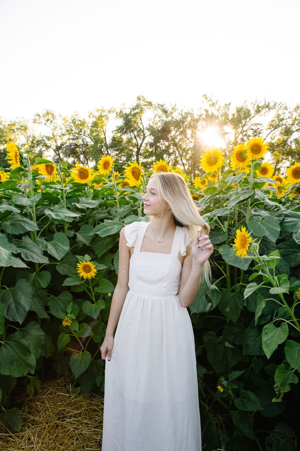 A white dress flows gracefully against a backdrop of tall blooming sunflowers at sunset.