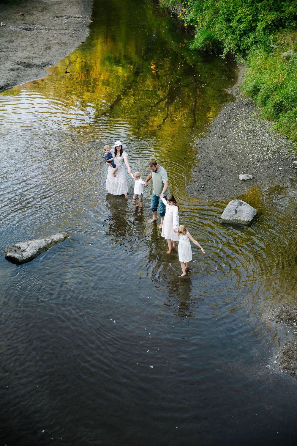 Group wading through shallow river water on sunny day surrounded by greenery.
