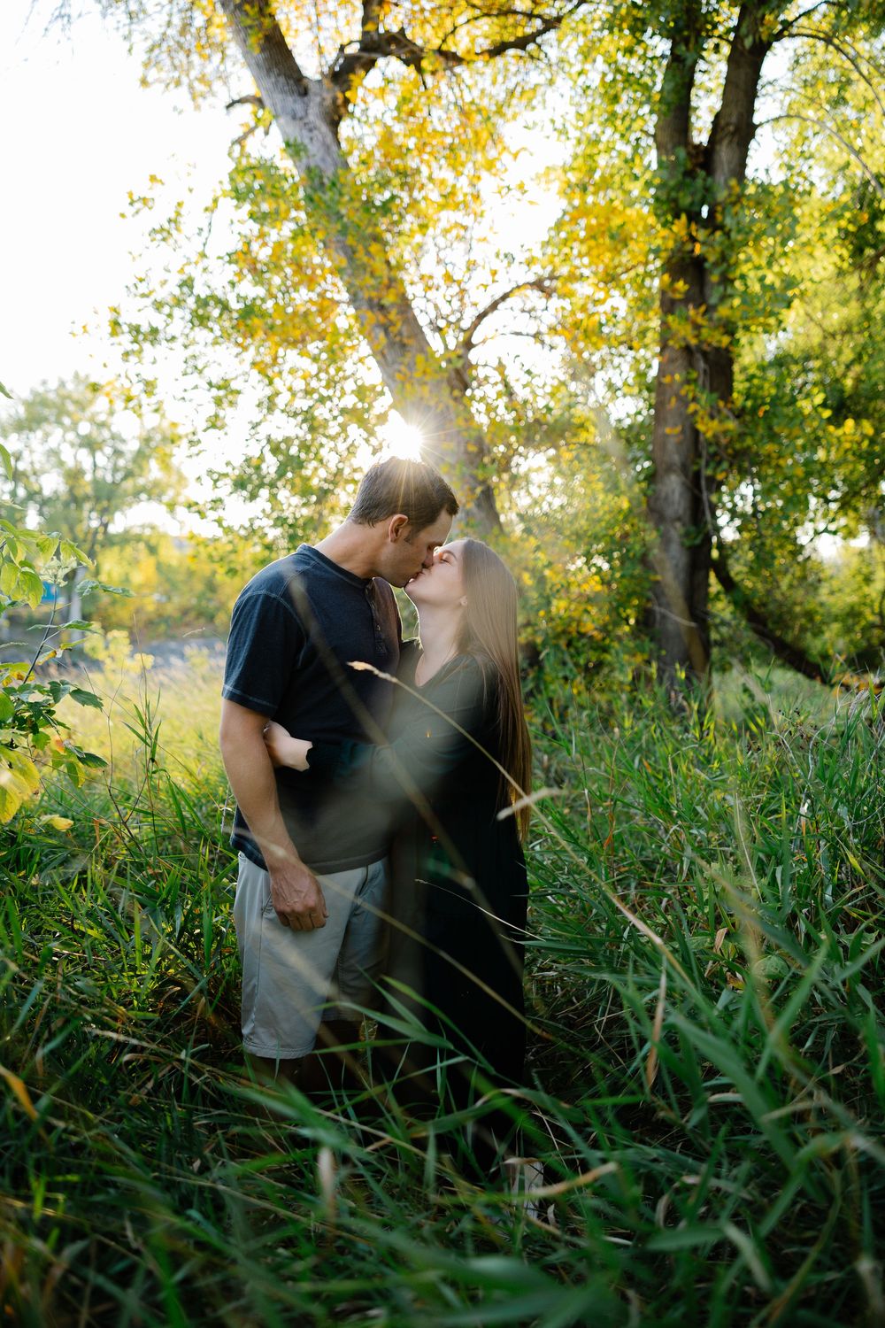 A couple shares an intimate moment in a grassy field during golden hour with sunlight filtering through autumn trees.