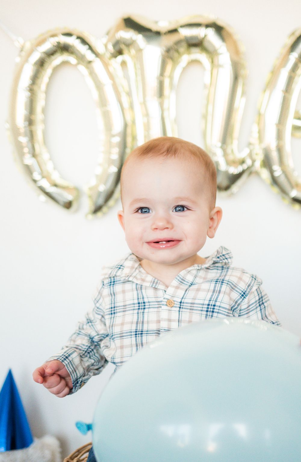 Smiling baby in plaid shirt poses with silver balloon numbers in background.