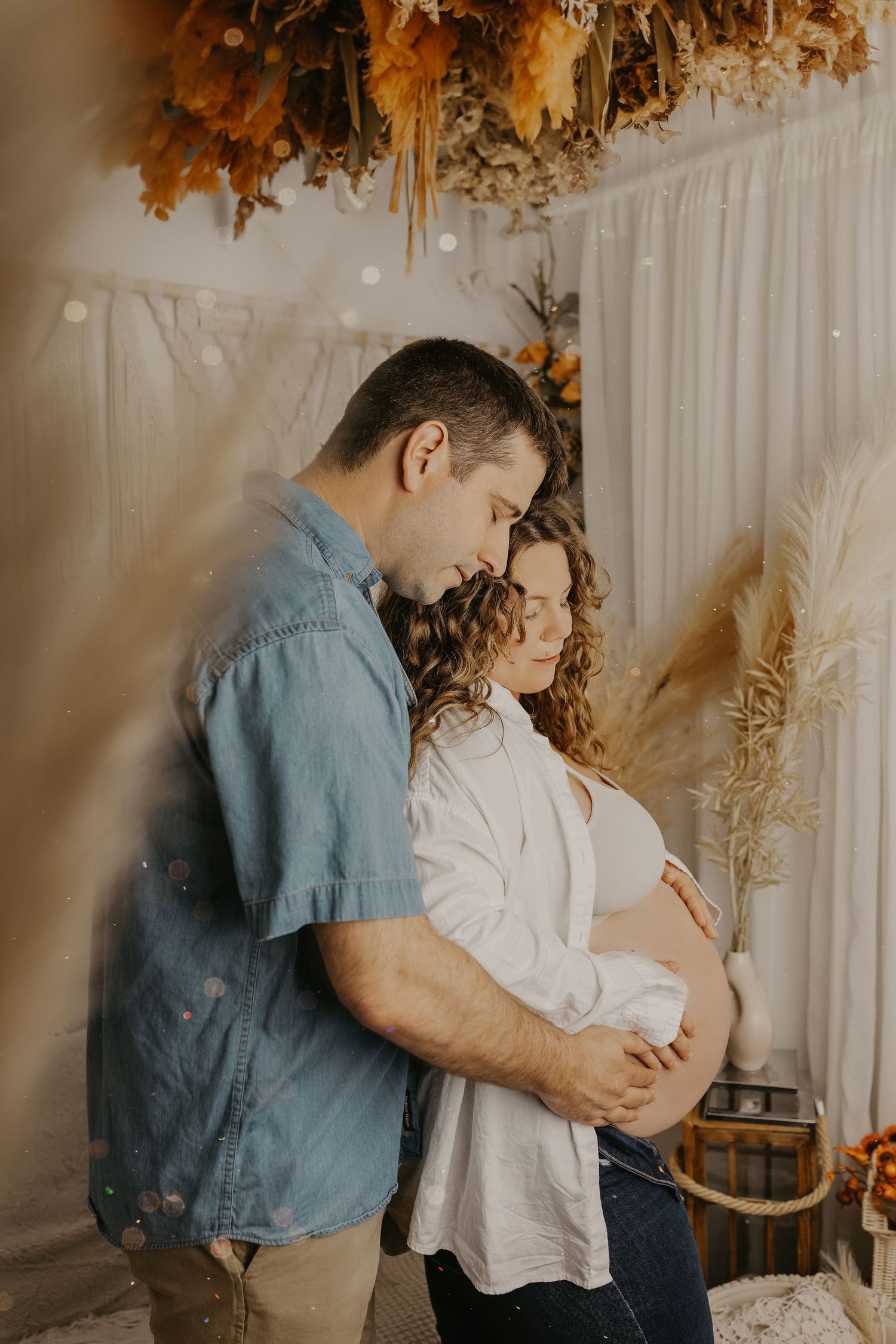 Intimate maternity photo of couple embracing with dried floral decor and warm lighting.