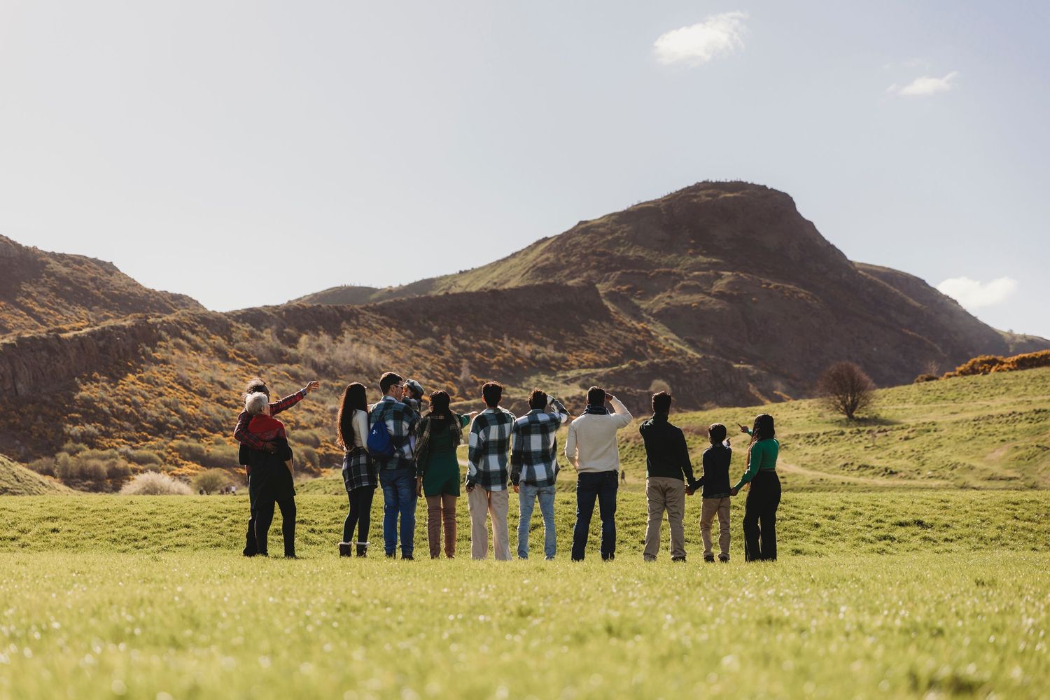 A group of people stand silhouetted against dramatic mountain scenery on a grassy field.