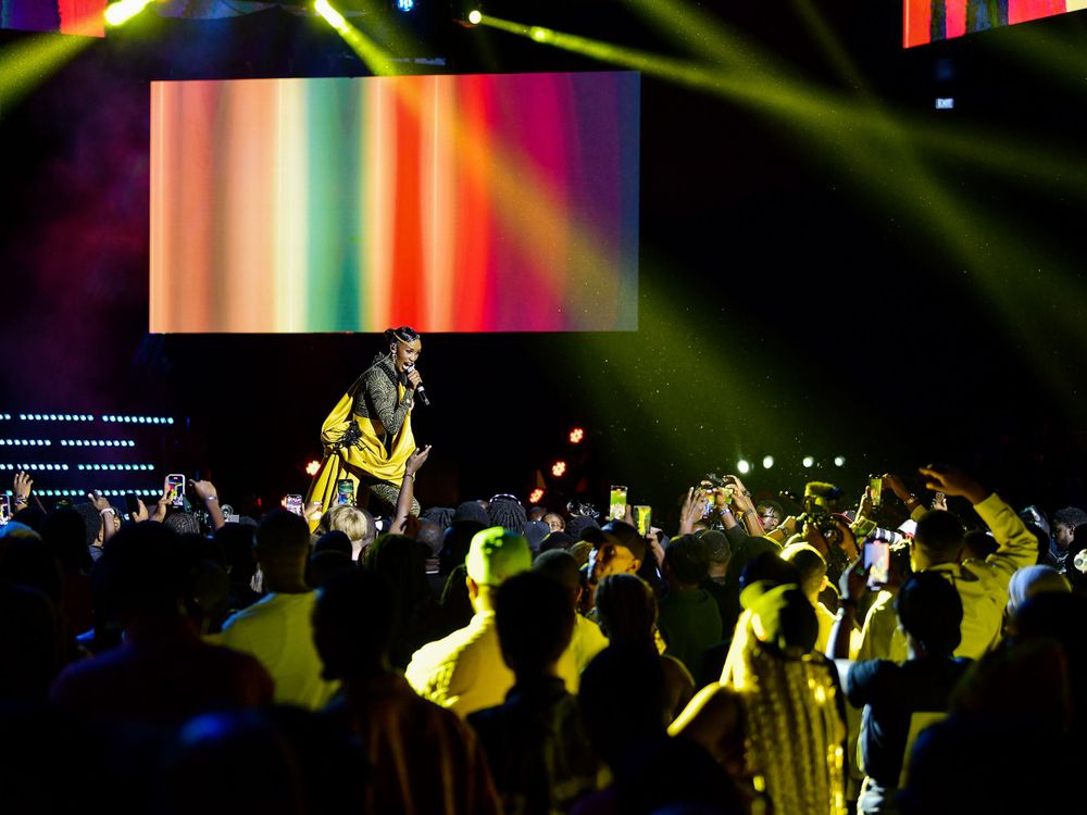 Concert performer in yellow outfit energetically entertaining crowd under colorful stage lighting and visual effects.
