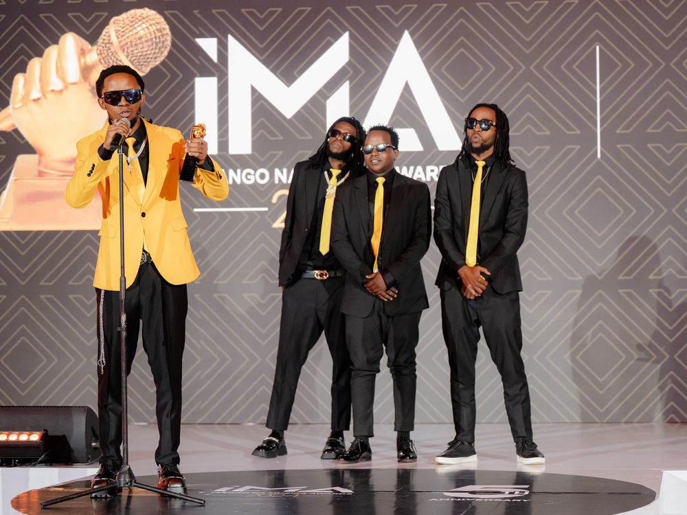 Group in coordinated outfits poses against IMA Awards backdrop.