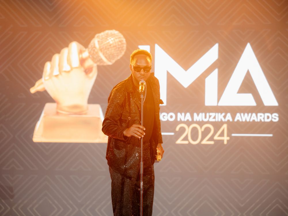 Solo performer speaks at microphone against illuminated IMA 2024 backdrop.