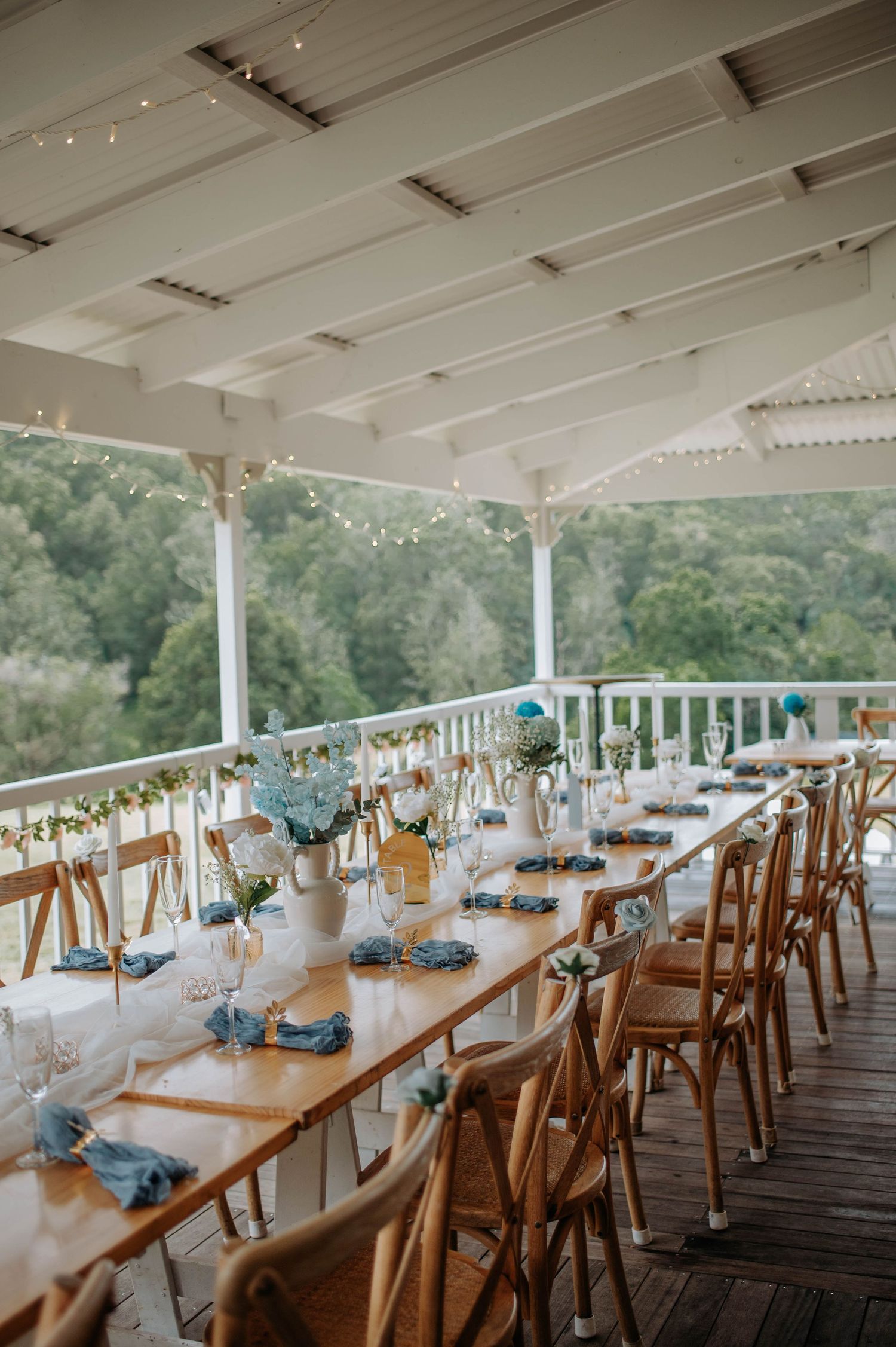 A rustic outdoor wedding reception table set with blue accents and wooden chairs on a covered porch.