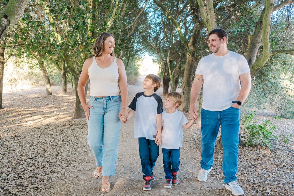 A family walks together on a dirt path through a wooded area on a sunny day.