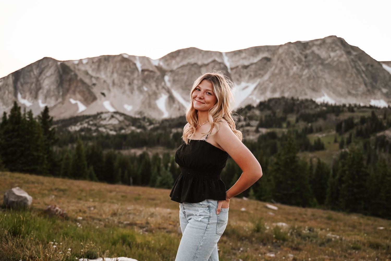 Woman in black top and jeans poses against mountain backdrop.