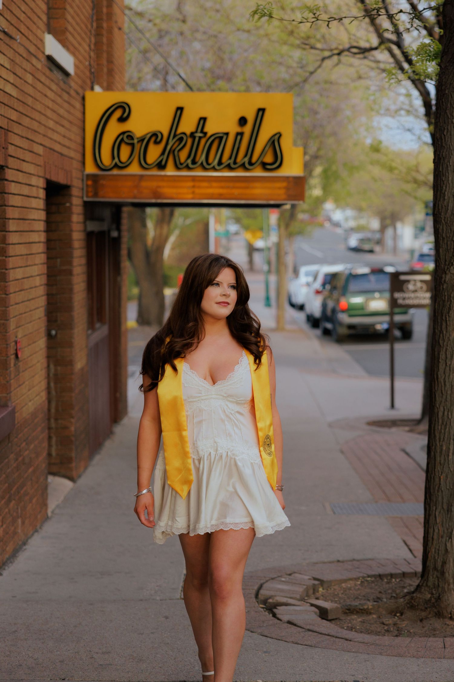 A brunette woman in a white dress and golden NAU graduation stole walks confidently under a Cocktails sign in downtown Flagstaff.