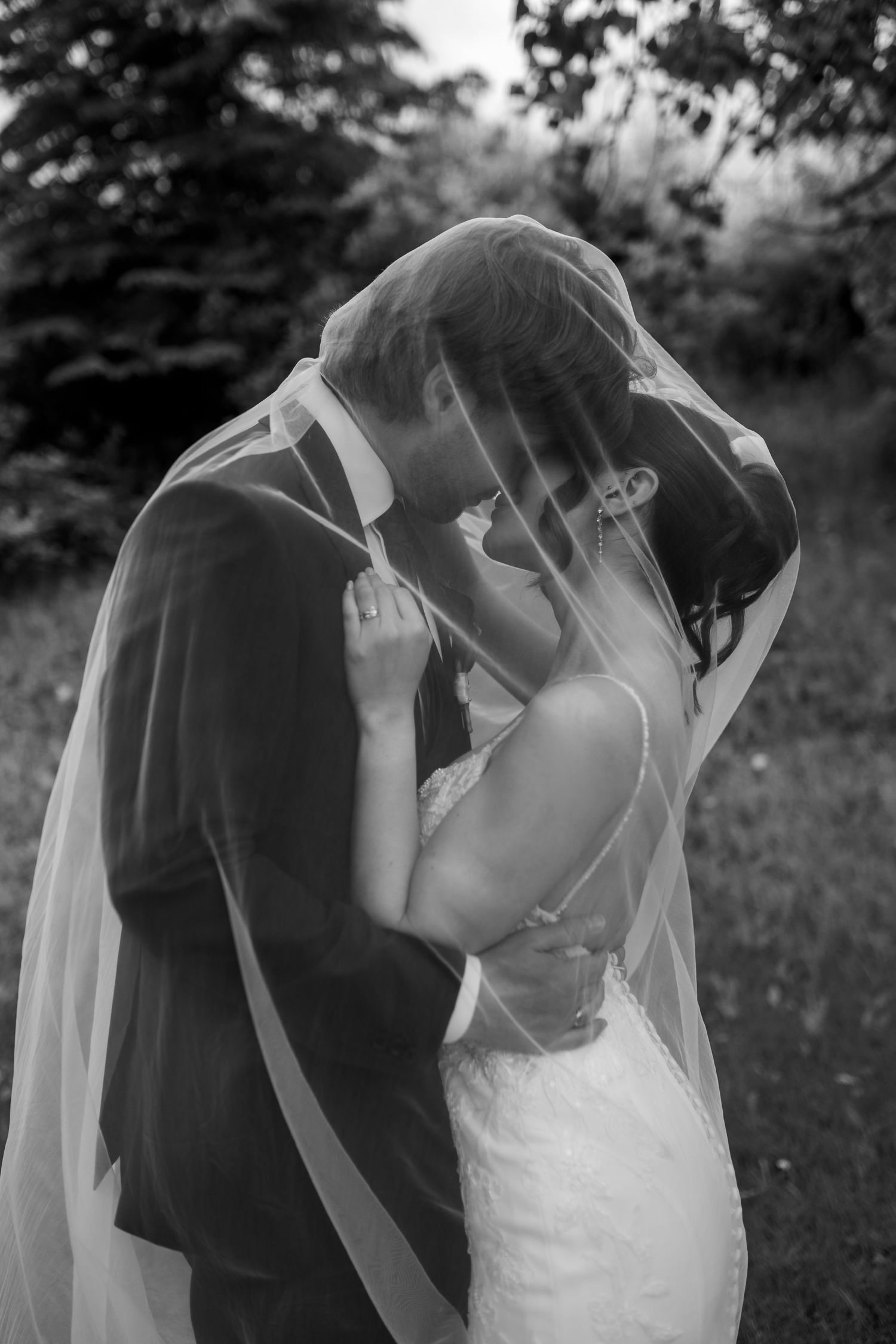Black and white photo of a bride and groom embracing intimately beneath a sheer wedding veil in an outdoor garden setting.