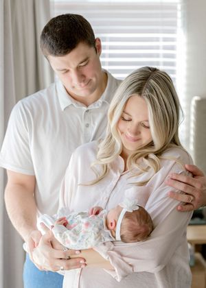 A family in white clothing shares tender moments with their newborn baby in a bright, sunlit room.