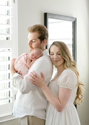 A couple in white outfits share tender moments with their newborn baby in a bright, sunlit room.