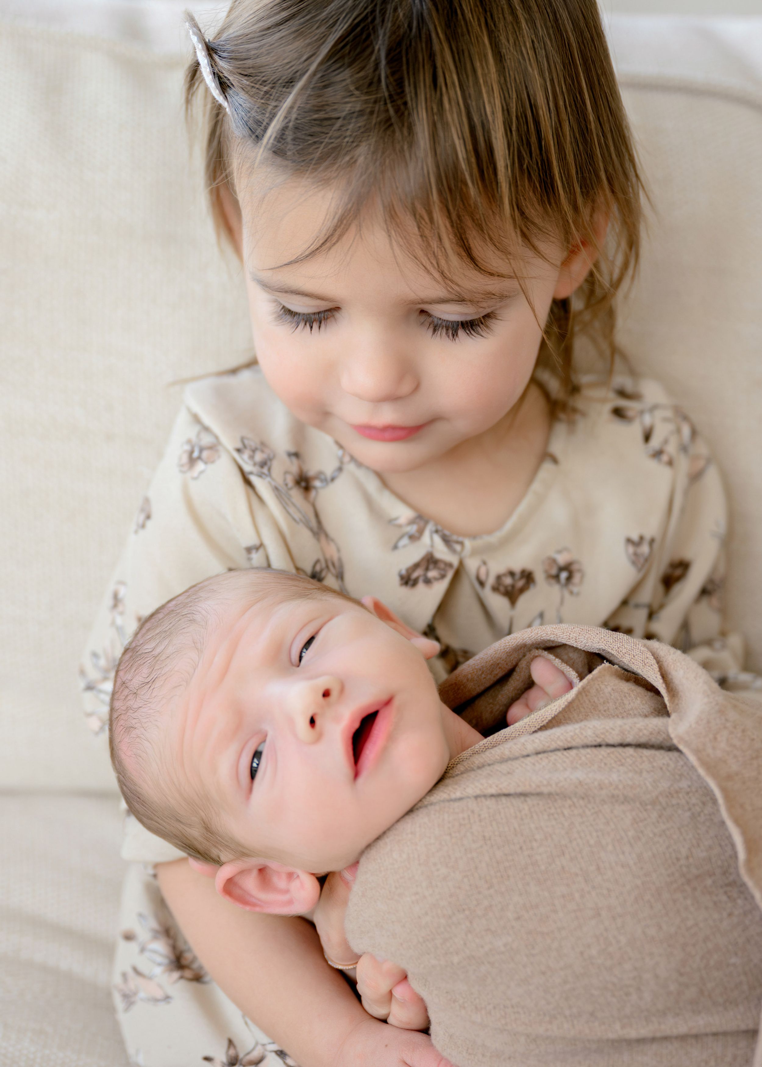A young child in beige clothing gently embraces and interacts with a sleeping newborn baby.