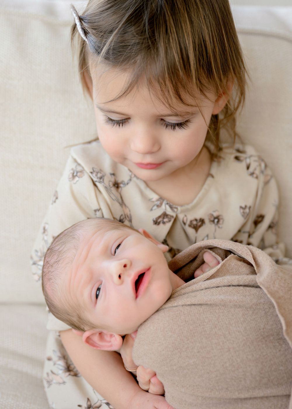 A young child in beige clothing gently embraces and interacts with a sleeping newborn baby.
