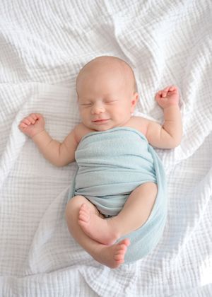 A sleeping baby wrapped in a light blue swaddle lies peacefully on white textured bedding.