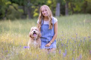A woman in a blue dress sits in a wildflower meadow with a small white dog during a sunny summer day.