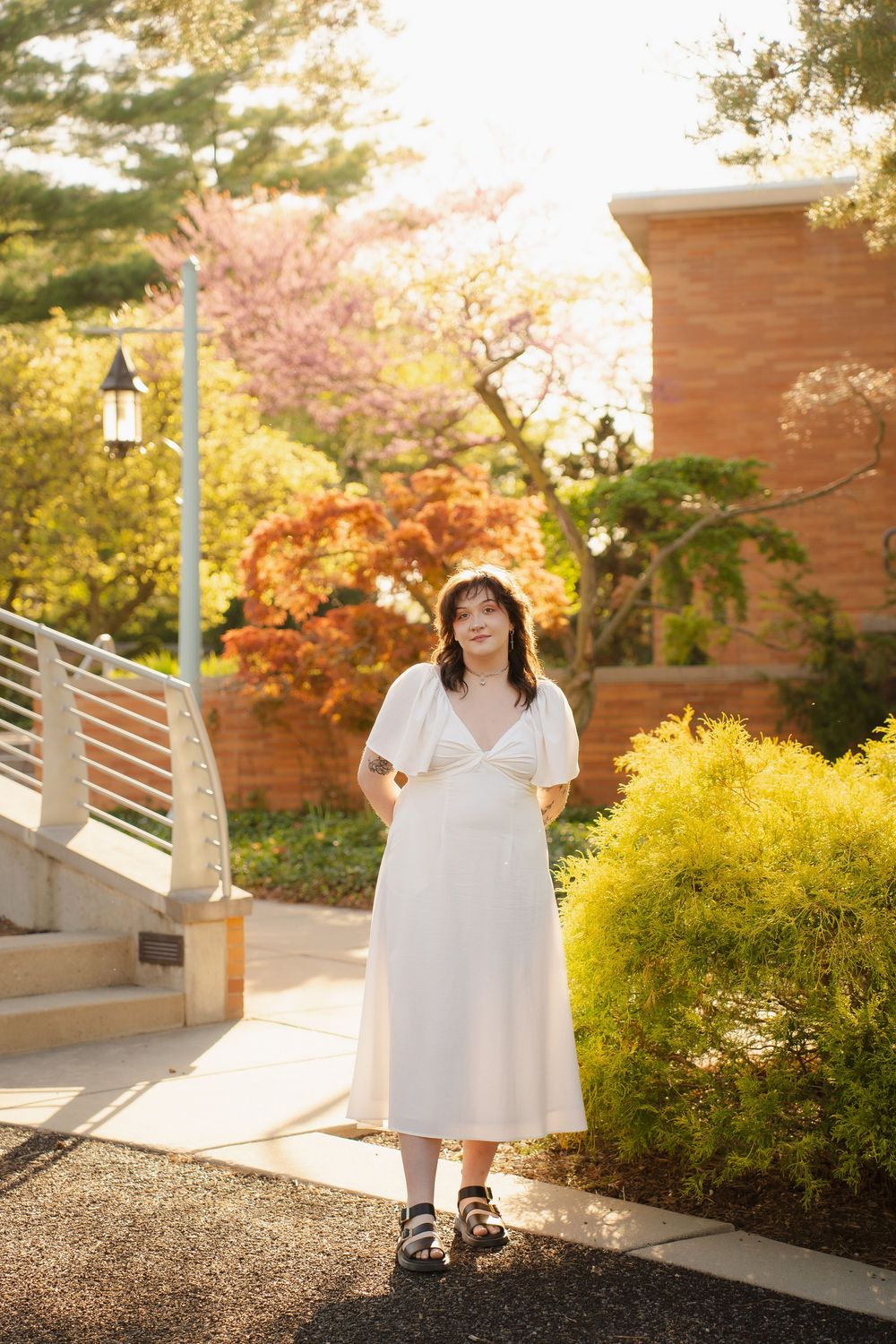 Woman in white midi dress with puff sleeves stands outdoors near brick building surrounded by colorful spring foliage.