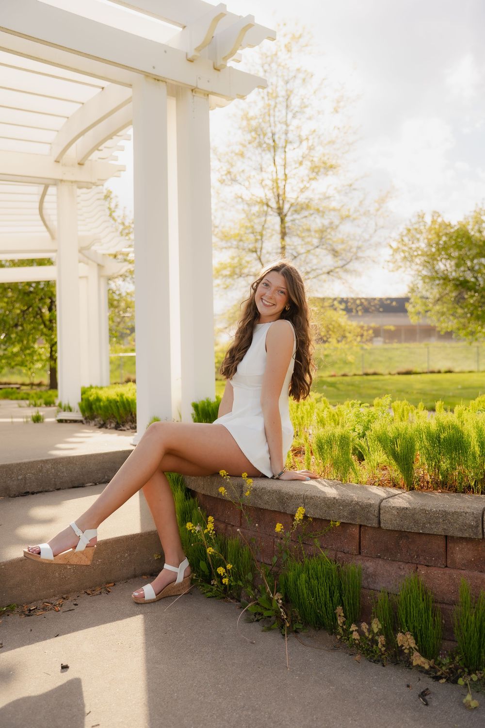 Young woman in white dress sitting on brick garden wall under white pergola, smiling outdoors in sunny park setting.