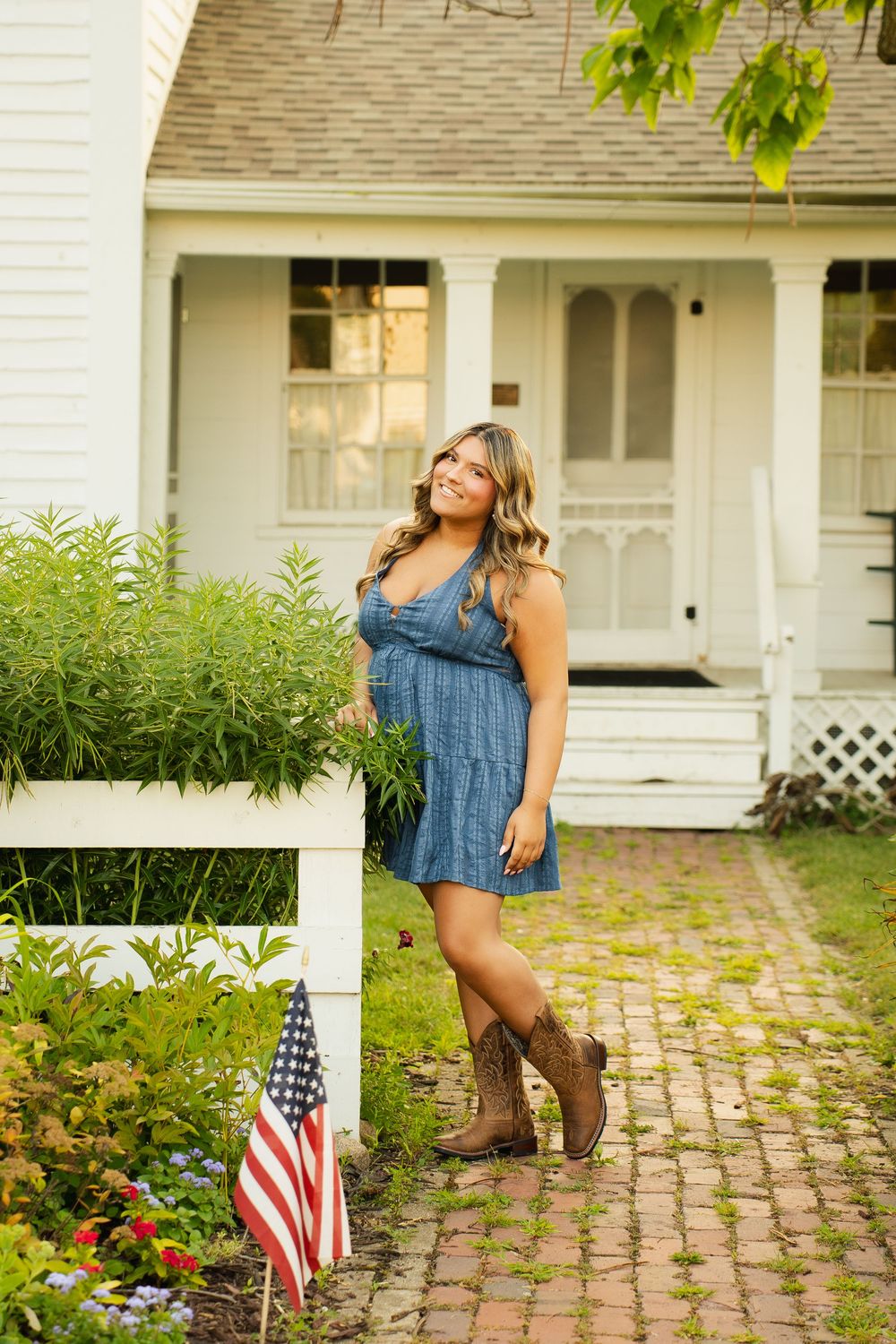 Young woman in denim dress and cowboy boots poses by white fence with American flag near charming white farmhouse.