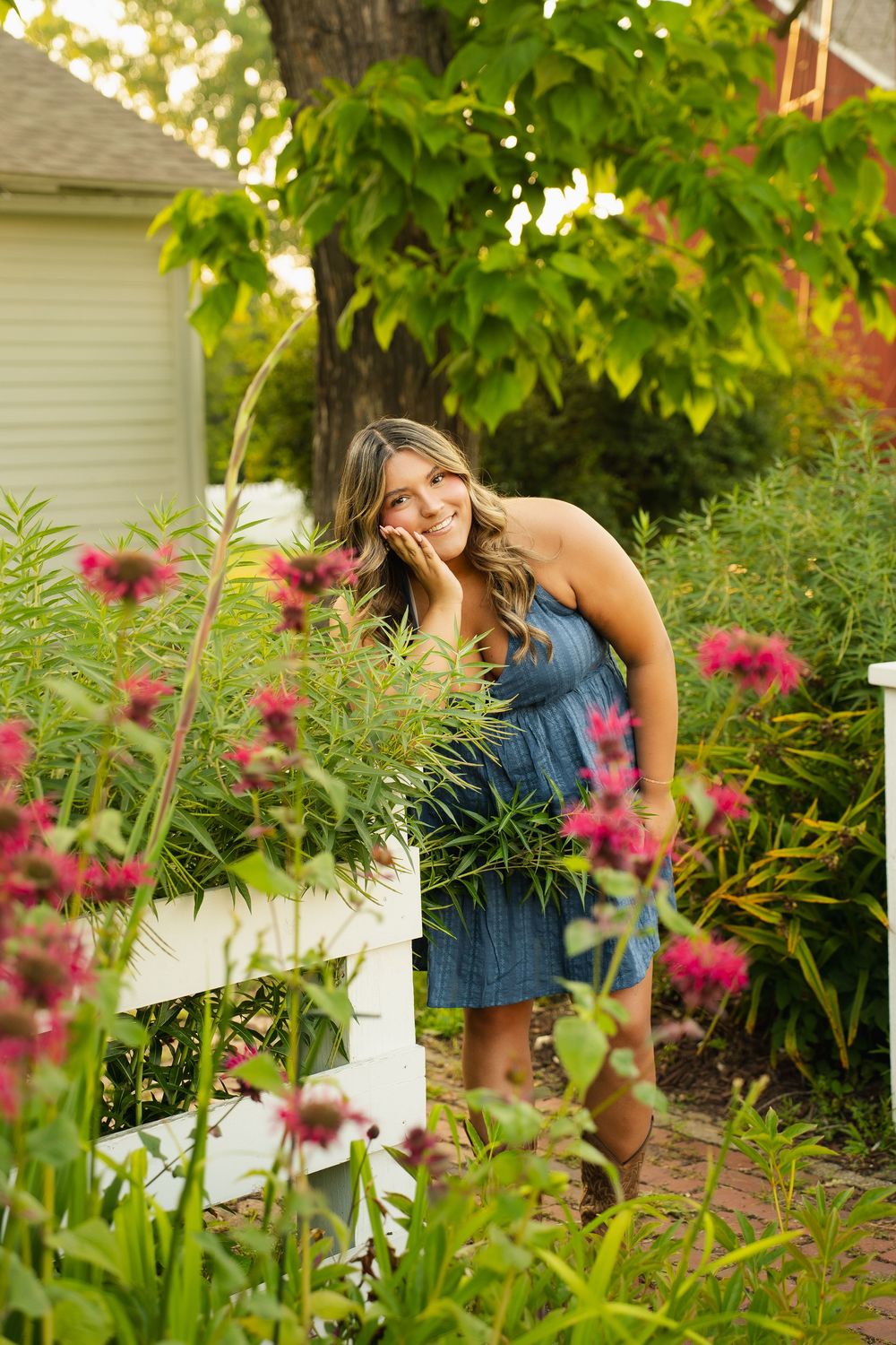 Young woman in denim dress smiling among colorful garden flowers by white fence.