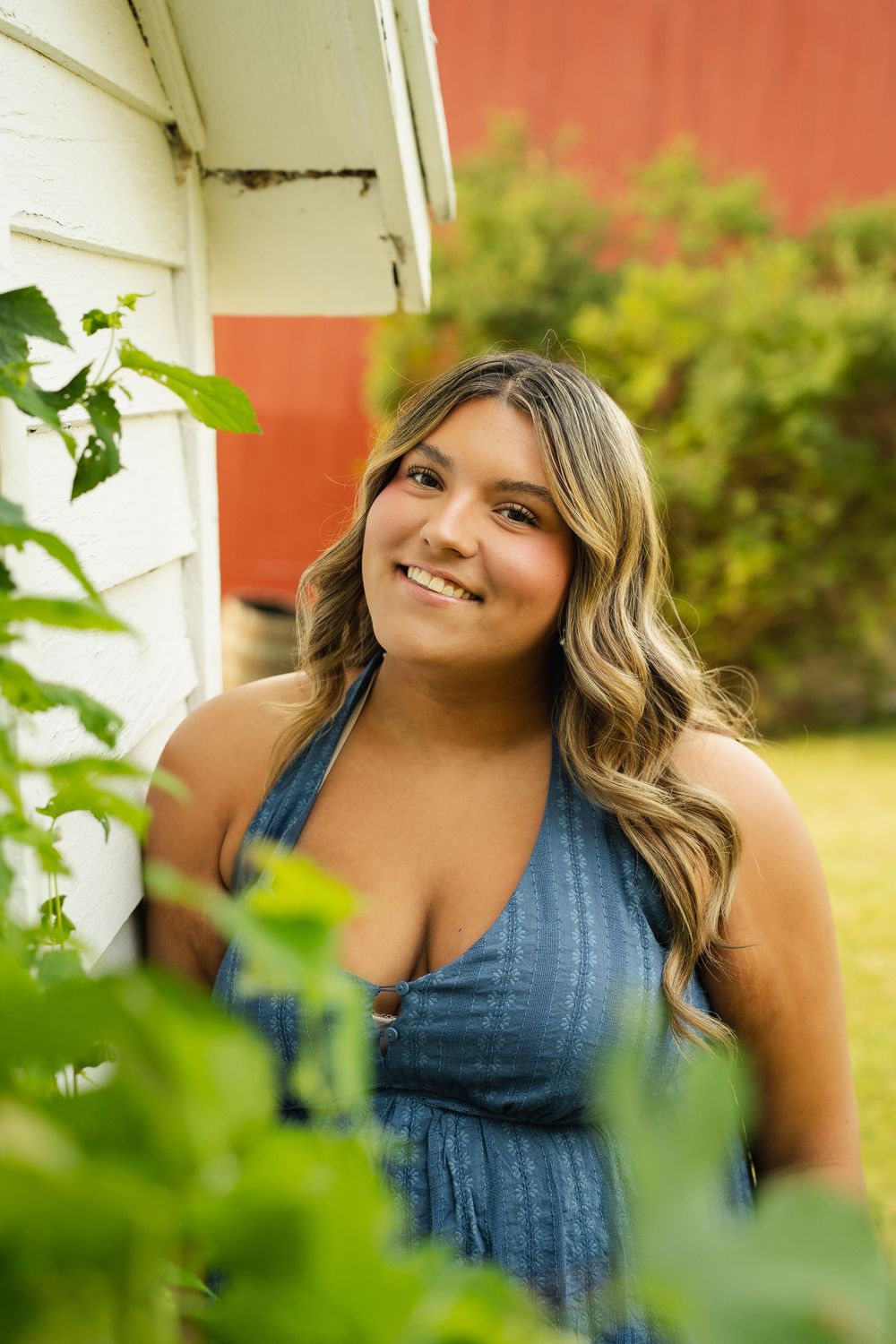 Smiling young woman with wavy blonde hair wearing a blue halter dress, posing outdoors near a white barn wall.