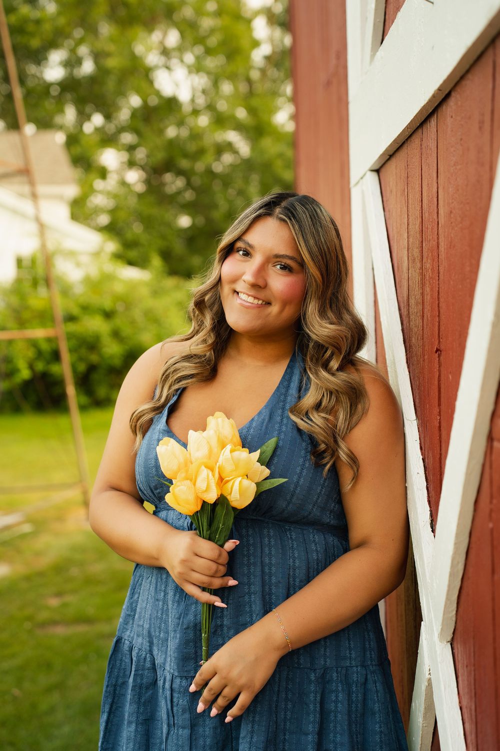 Smiling young woman in blue dress holding yellow tulips leaning against red barn outdoors.