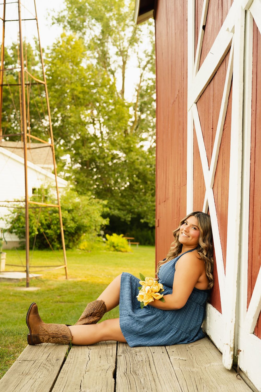 Young woman in blue dress and cowboy boots sits against red barn holding yellow flowers, smiling outdoors.