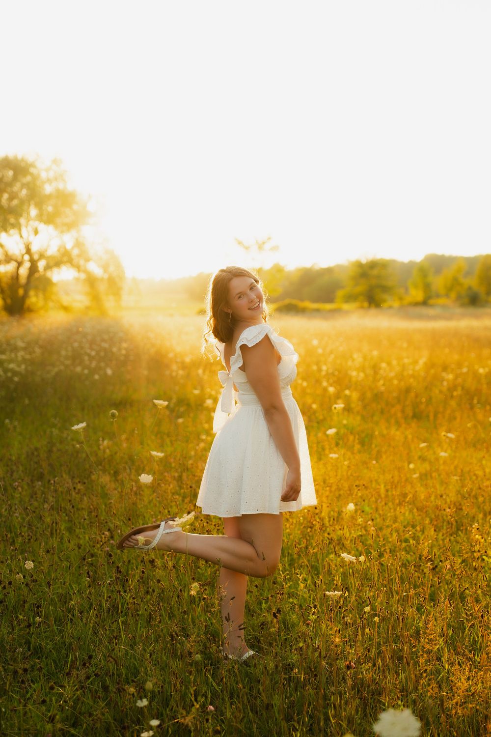 Young woman in white dress standing in golden sunlit field at sunset, smiling over shoulder with one leg raised.