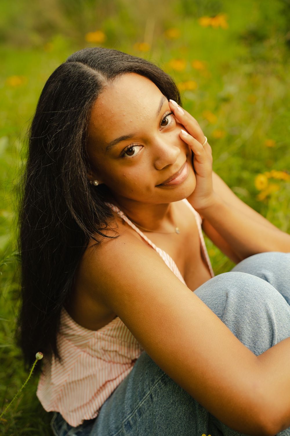 Young Black woman with long hair sits in a green meadow with yellow wildflowers, smiling softly with hand on cheek.