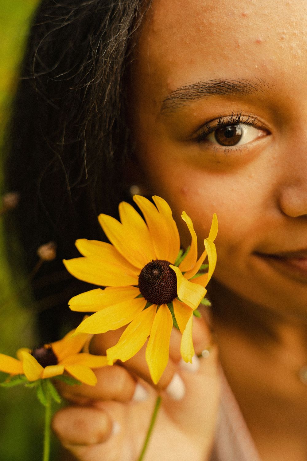 Young woman holding a bright yellow black-eyed Susan flower close to her face, smiling softly outdoors.
