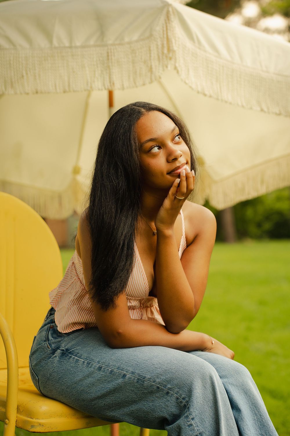 Young Black woman sitting outdoors on yellow chair, resting chin on hand, wearing jeans and tank top near fringed umbrella.