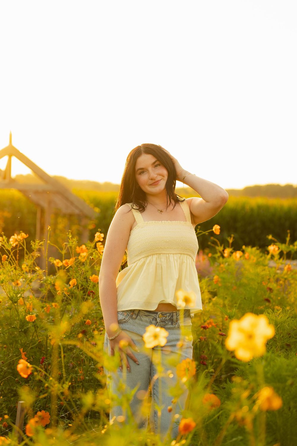 Young woman in yellow top and jeans poses among wildflowers in golden sunset light.