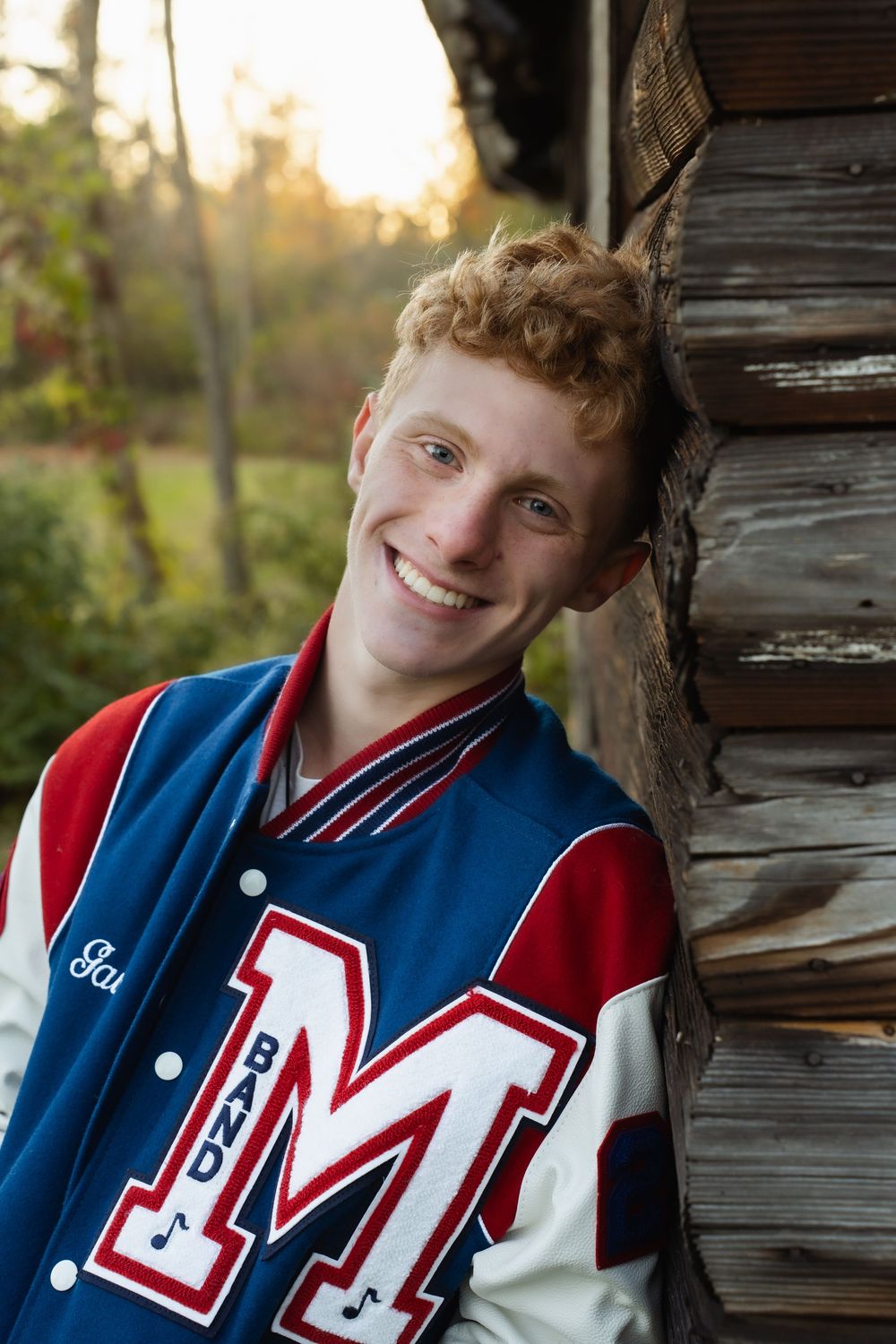 A high school senior wearing a varsity letterman jacket poses against rustic wooden boards at sunset.