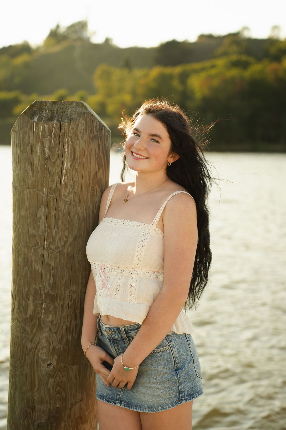 Young woman in white crop top and denim skirt smiling by wooden post near river at golden hour.