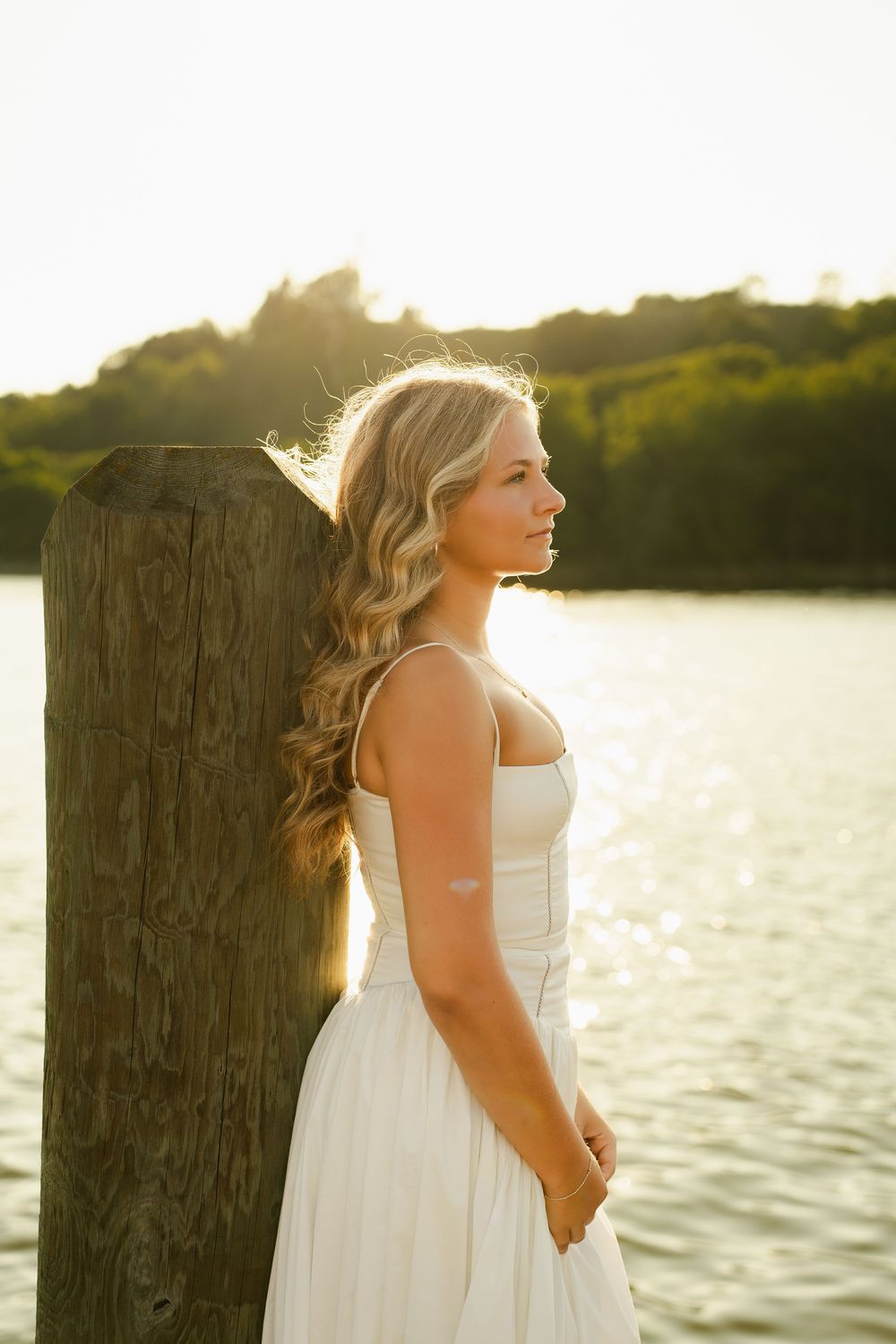 Young blonde woman in white dress stands by wooden post at golden hour sunset near serene water.