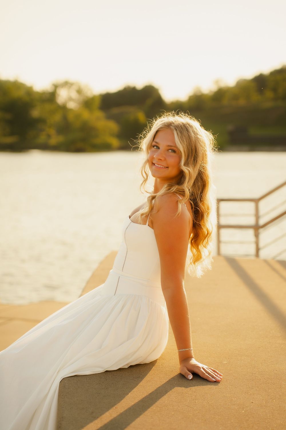Young blonde woman in white dress sitting on a dock at golden hour sunset by a lake, smiling over her shoulder.