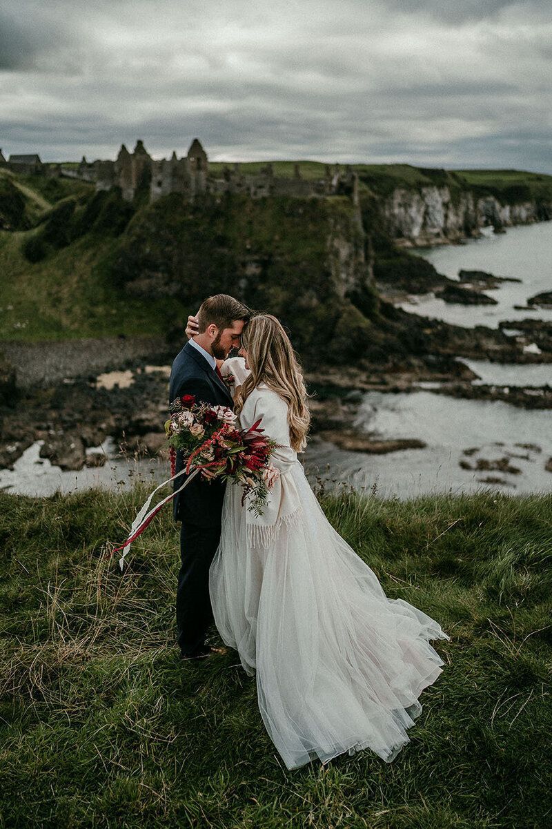 Bride and groom embracing on coastal cliffs with Dunluce Castle ruins in the background.