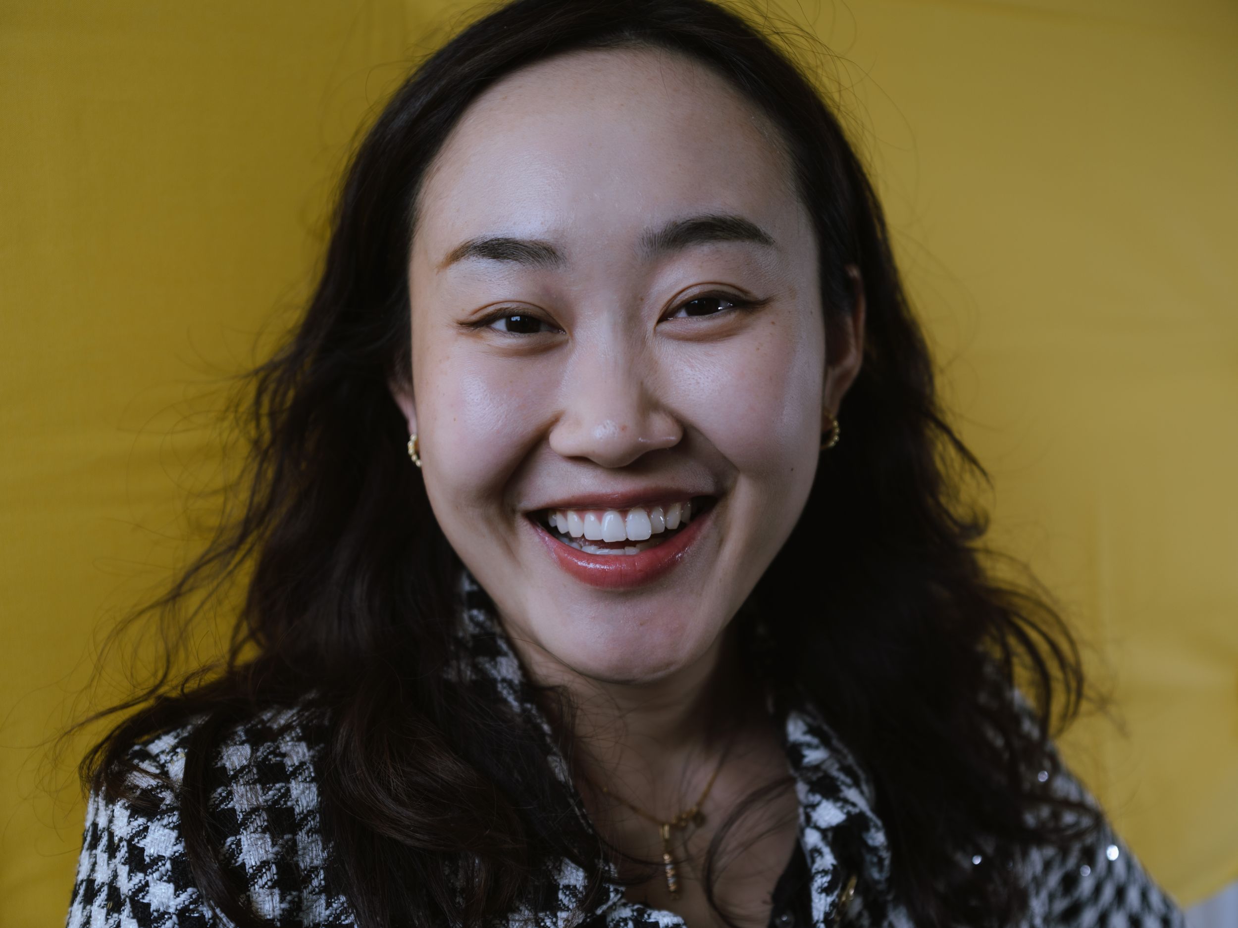Asian woman with long dark hair laughing joyfully wearing a houndstooth blazer against a yellow background.