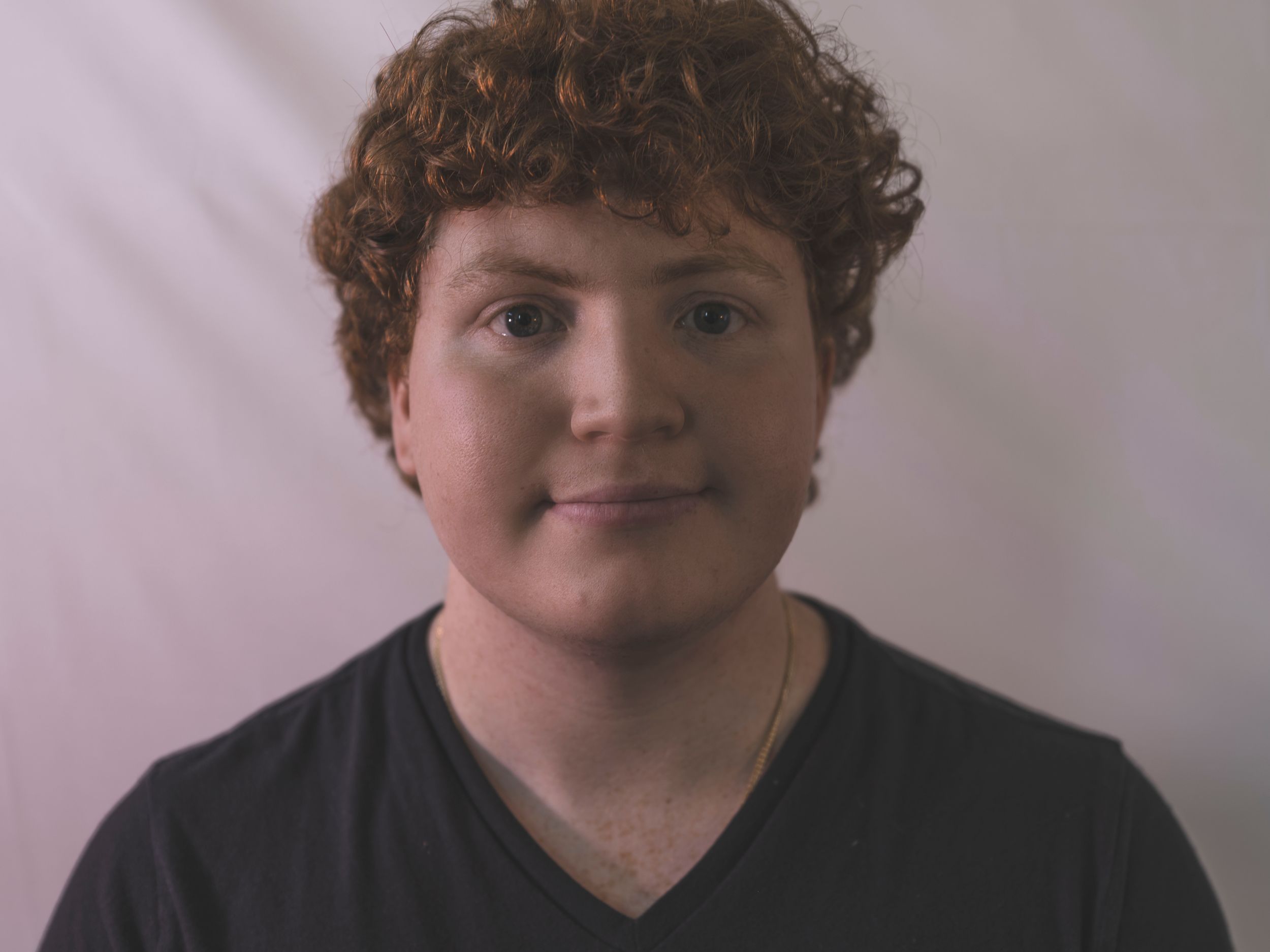 Young person with curly red hair wearing a black v-neck shirt smiling against a white background.